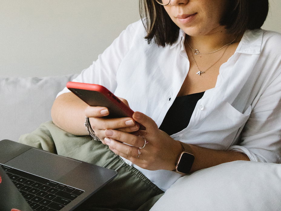Eine Frau mit Laptop auf dem Schoß checkt ihr Smartphone.